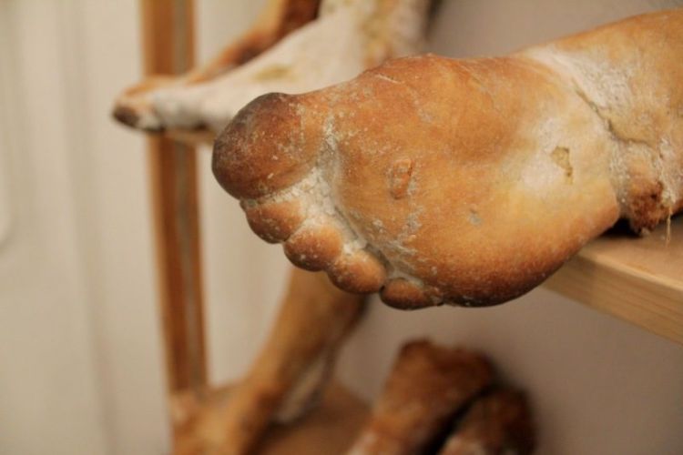 Kelt tésztából mintázott emberi lábfejek, 3 sorban fa polcokon a falhoz közel.